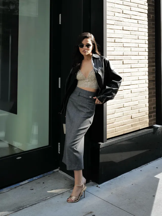 Rachel Zegler wearing a black jacket, grey skirt and pale heels outdoors.