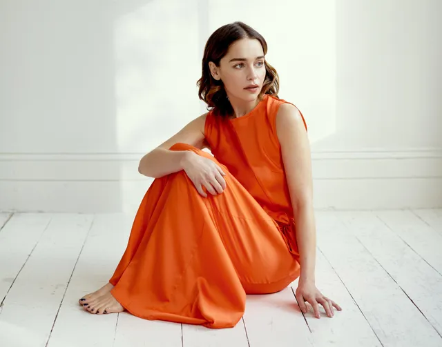Emilia Clarke sitting barefoot on a white floor in an orange dress.