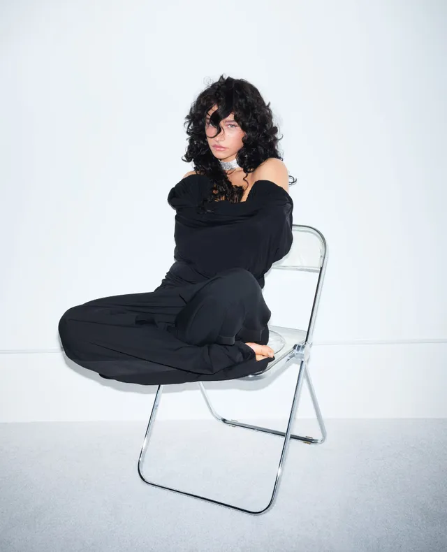Odessa A'zion sitting barefoot cross-legged on a chair.
