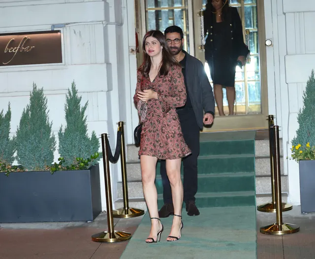 Alexandra Daddario standing outside in a patterned dress and heels.
