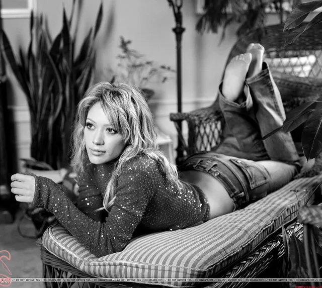 Hilary Duff lying on a chaise longue wearing heeled shoes in a black and white portrait.