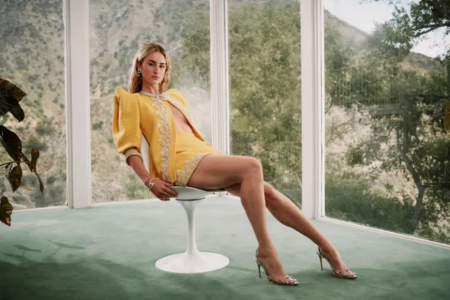 Grace Van Patten sitting by a window in a yellow outfit and pointed heels.