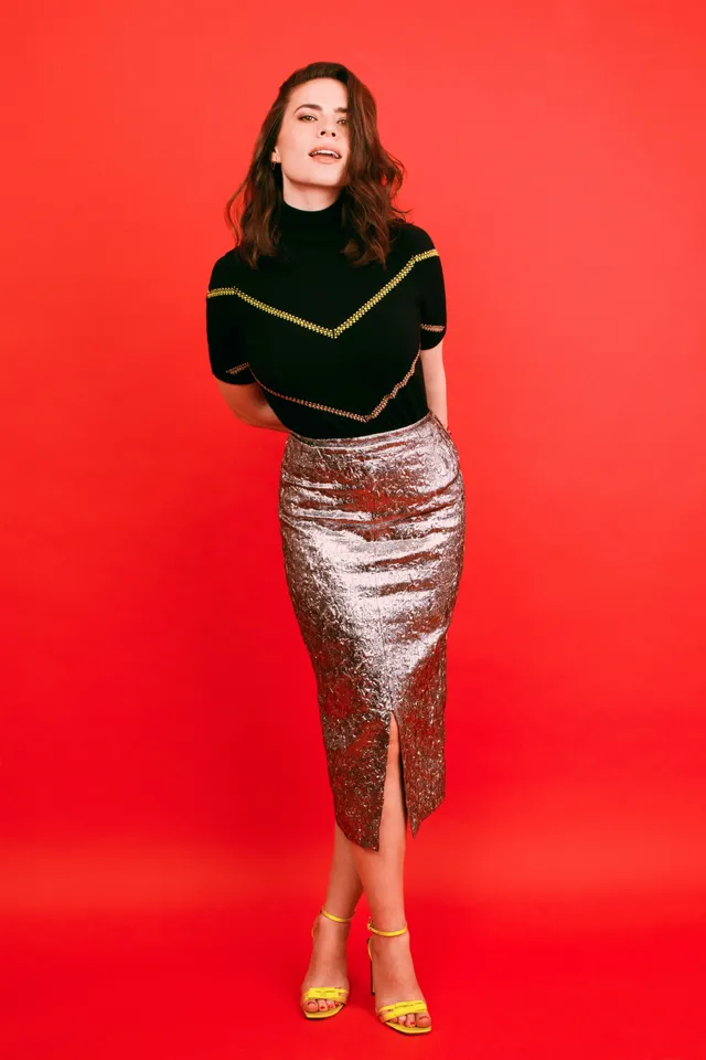 Hayley Atwell posing against a red wall in a patterned skirt.