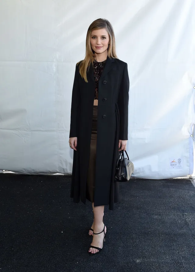 Dianna Agron standing in a dark dress and long coat.