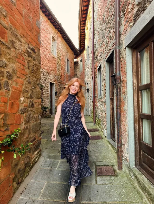 Isla Fisher walking along a stone lane in a patterned dress and wedge sandals.