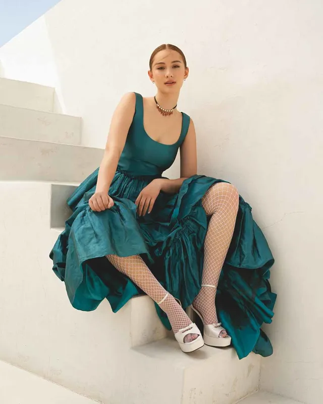 Lola Tung sitting on steps in a teal dress, light socks and white heels.