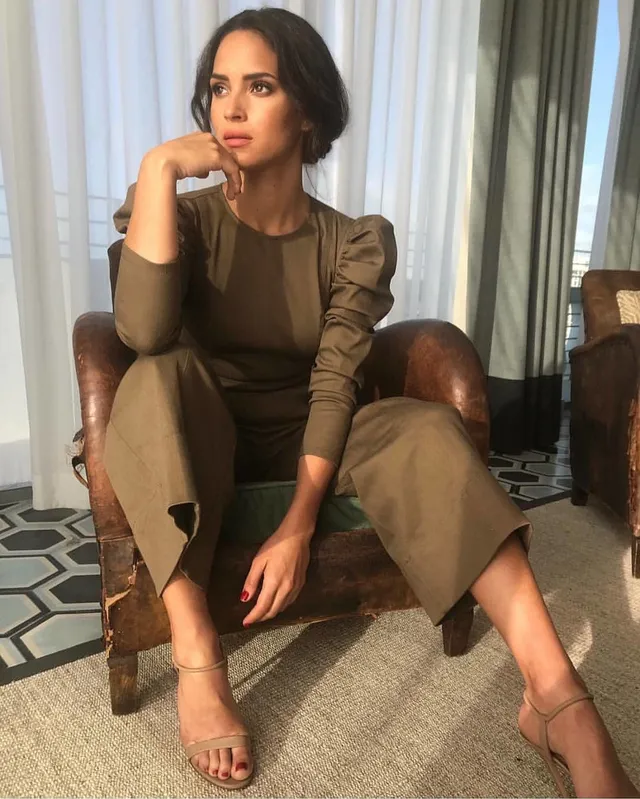 Adria Arjona sitting in an armchair wearing an olive outfit and heeled sandals.