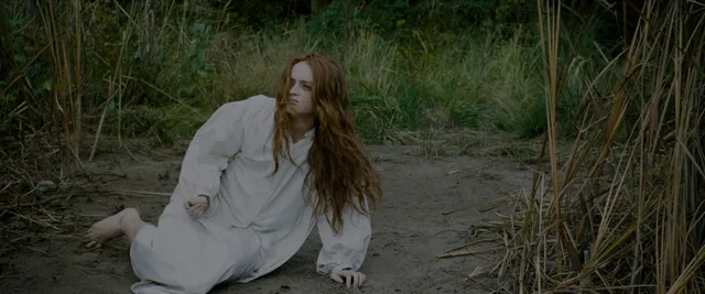 Sadie Sink sitting barefoot on a sandy path in a film scene.