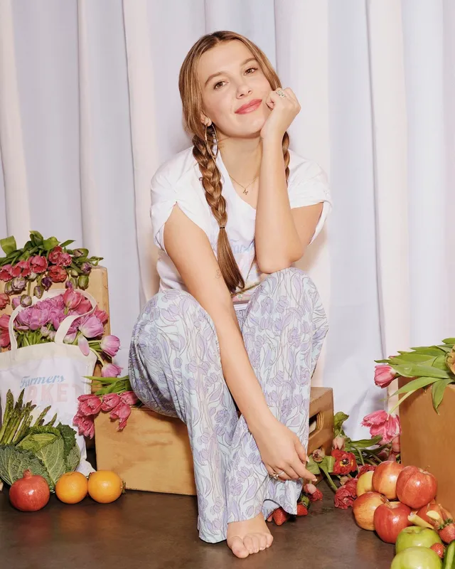 Millie Bobby Brown sitting barefoot among flowers and fruit for a studio portrait.