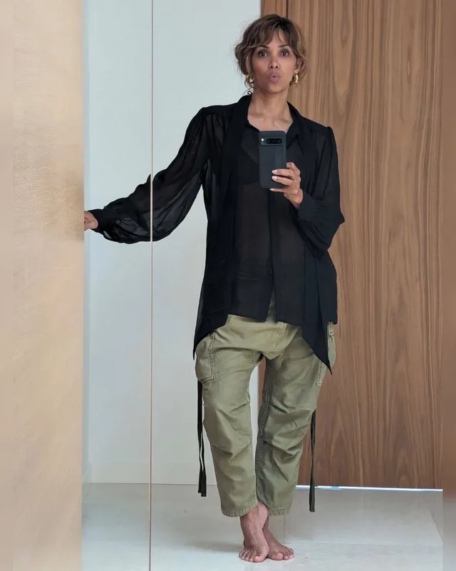 Halle Berry standing barefoot indoors in a black shirt and light trousers.