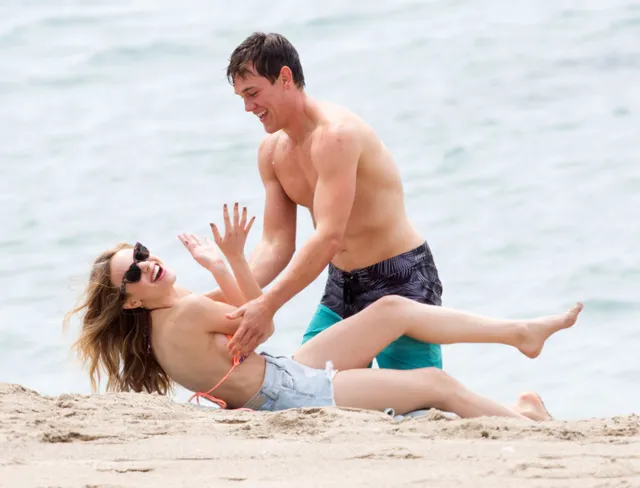 Halston Sage reclining barefoot on a beach beside a man.