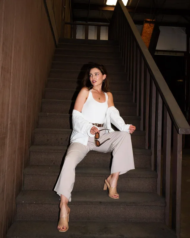Danielle Campbell sitting on stairs in a white blouse and pale trousers.