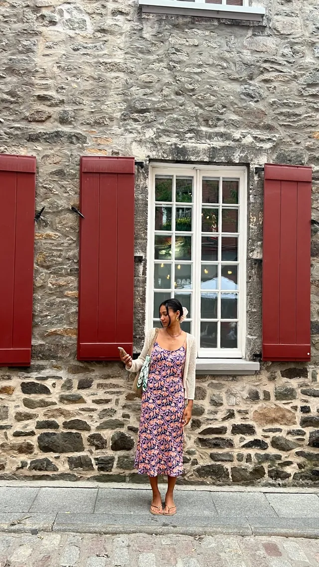 Greta Onieogou standing beside a stone wall in a floral dress and flats.