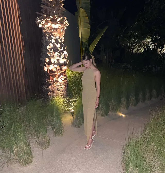 Halston Sage standing outdoors at night in a pale dress and strappy heels.