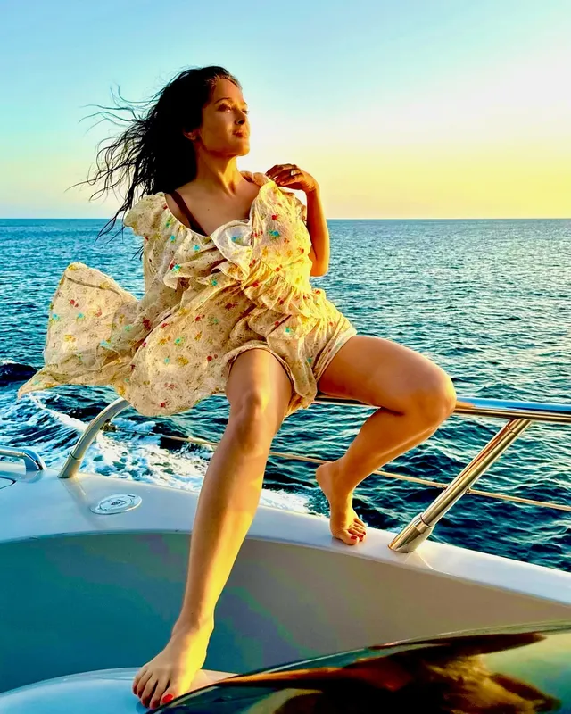Salma Hayek sitting barefoot on a boat railing at sunset.
