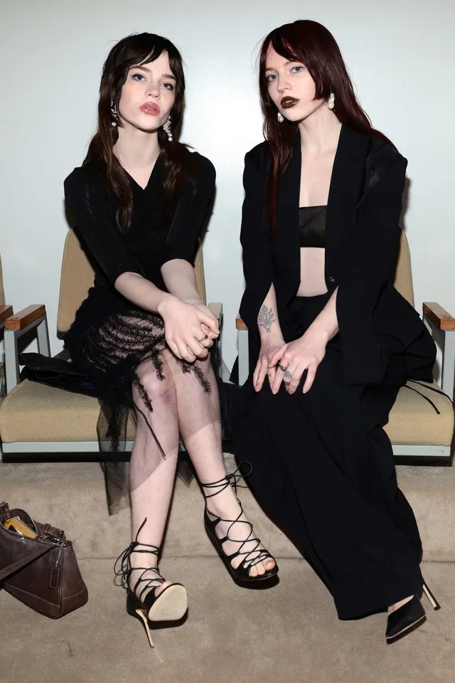Sophie Thatcher sitting beside another woman in a black dress and strappy heels.