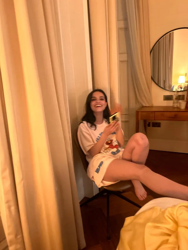 Rachel Zegler sitting barefoot on a chair beside a bed.