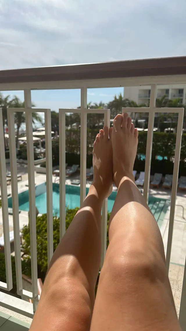 Paige Spiranac resting her bare feet beside a balcony railing.