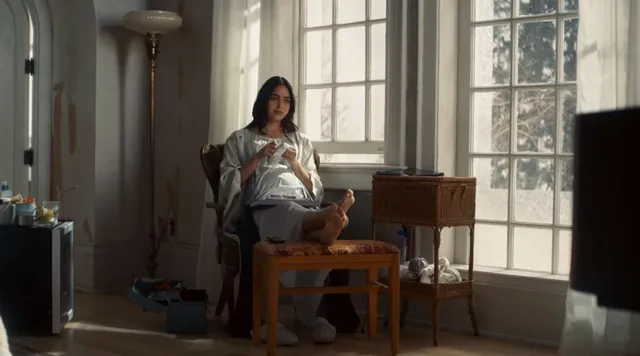 Melissa Barrera sitting barefoot in an armchair near a window.