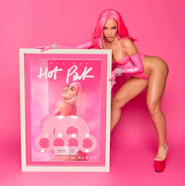 Doja Cat posing beside a framed album display in red platform heels.