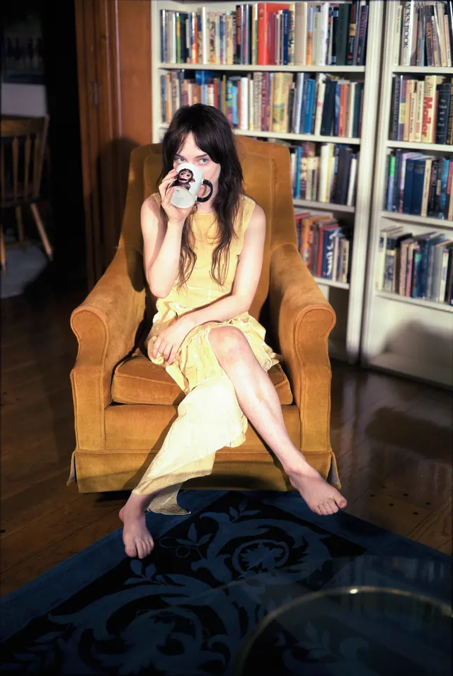Sophie Thatcher sitting barefoot in an armchair beside shelves of records.