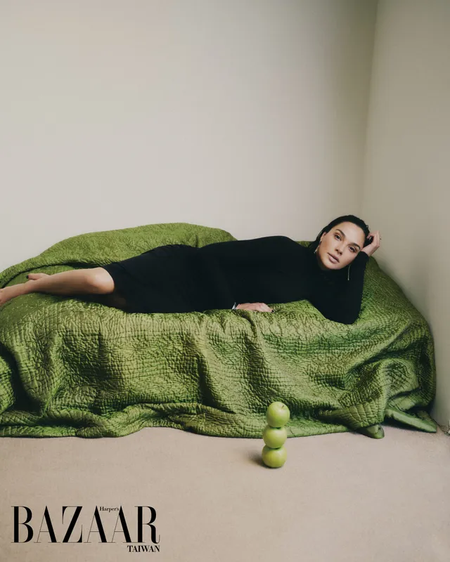 Gal Gadot reclining barefoot on a green sofa.