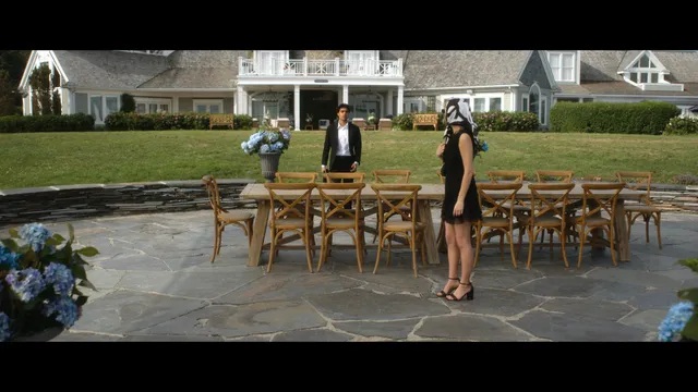 Emily Alyn Lind standing outdoors in a short black dress and strappy heels in a scene.