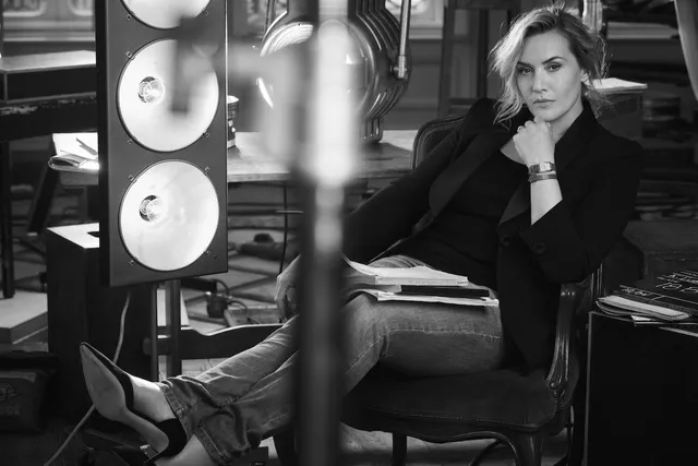 Kate Winslet sitting indoors in a dark outfit and boots.