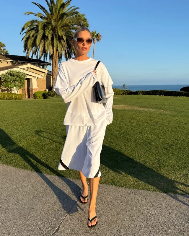 Elsa Hosk walking beside a lawn in a white outfit and black heeled sandals.