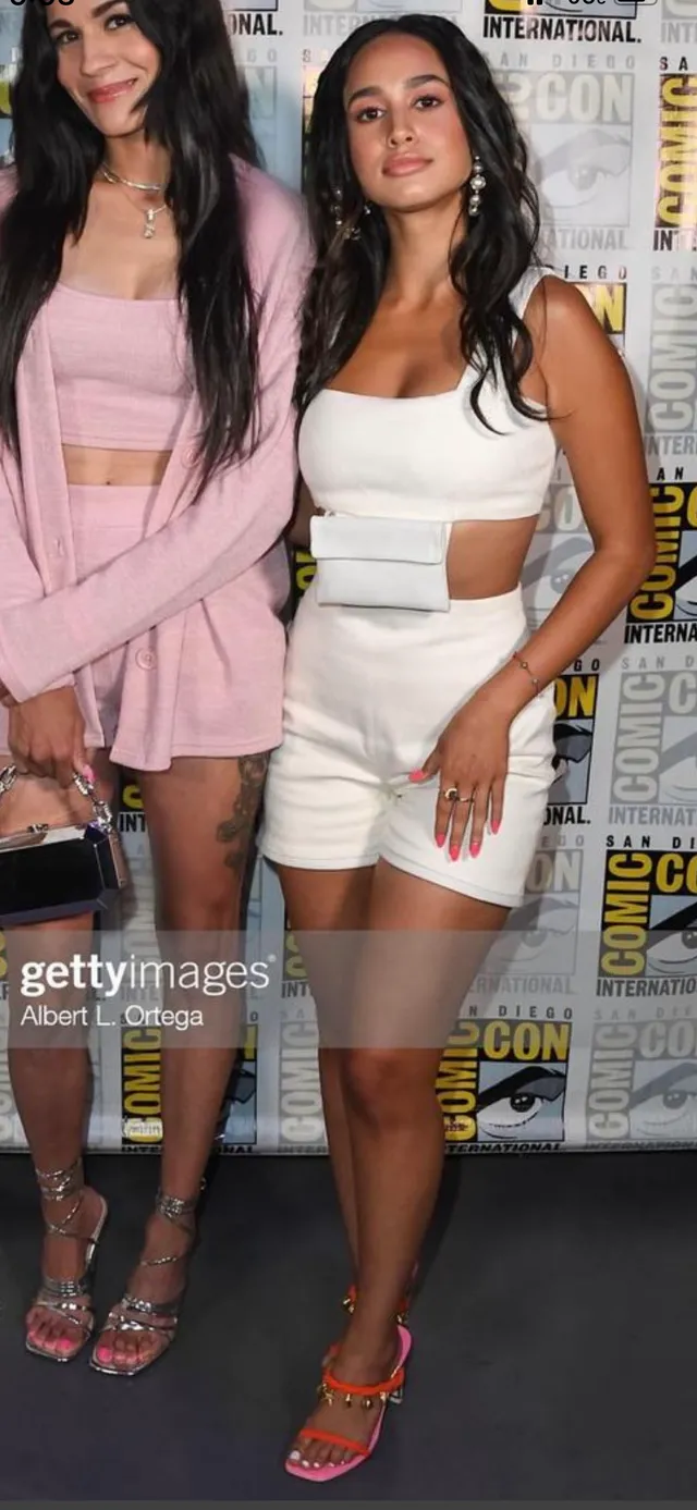 Emily Tosta posing at an event in a white outfit and orange heels.