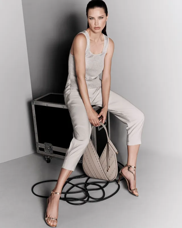 Adriana Lima sitting on a stool in a pale outfit and metallic heels.
