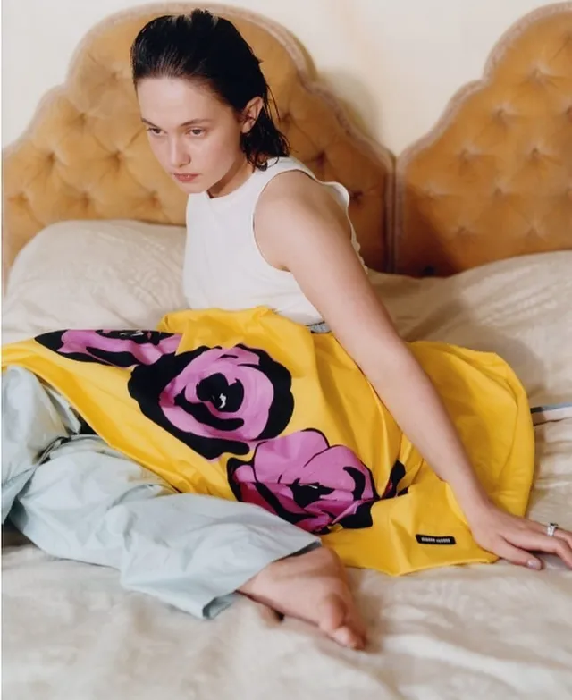 Cailee Spaeny sitting barefoot on a bed in a white top and bright skirt.