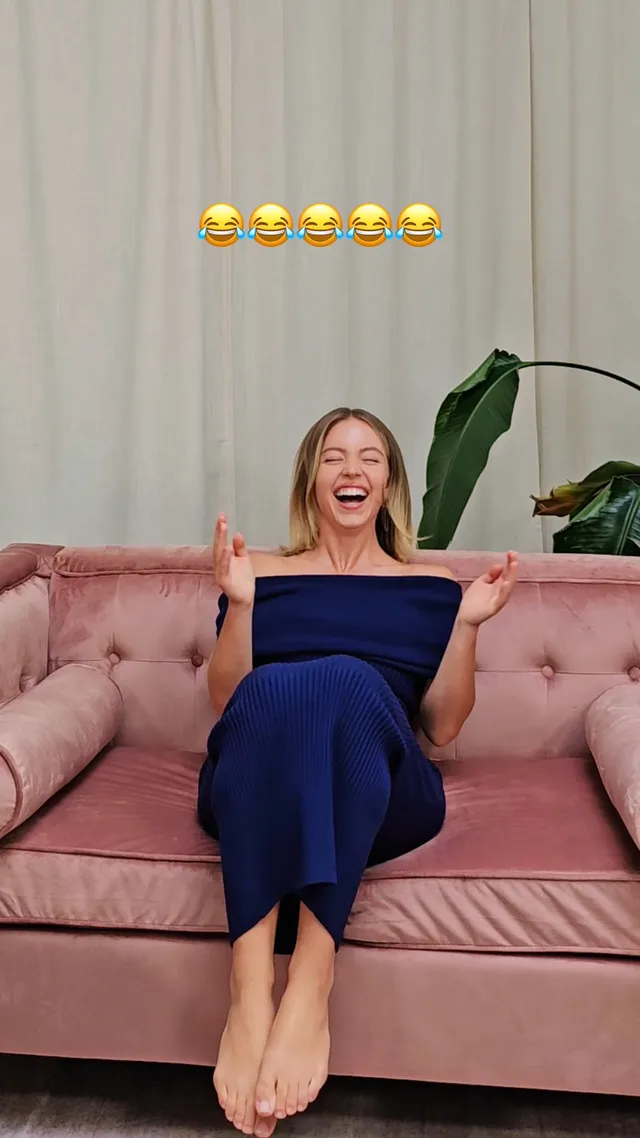 Sydney Sweeney sitting on a pink sofa in a blue outfit with her legs raised.