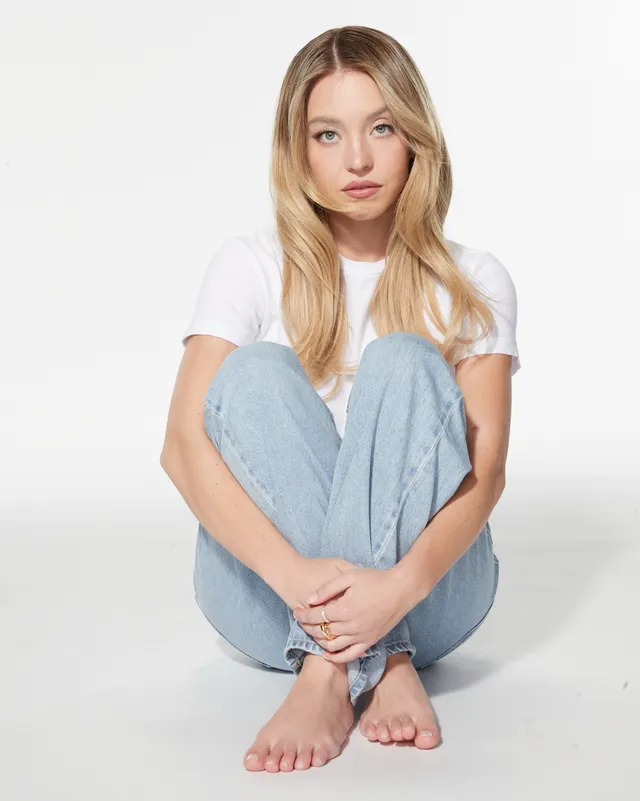 Sydney Sweeney: seated barefoot in jeans against a light background.