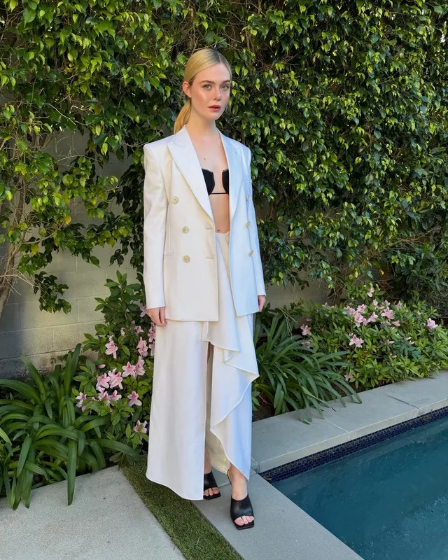 Elle Fanning wearing a white suit and black heeled sandals beside a pool.