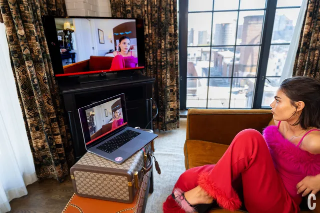 Ariana Greenblatt sitting indoors in a red outfit and flat sandals.
