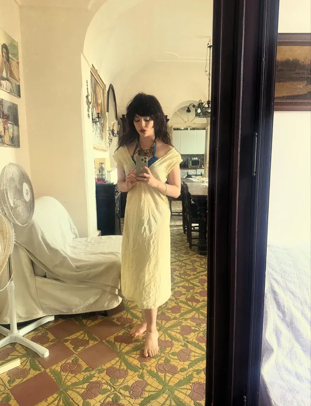 Sophie Thatcher standing barefoot in a yellow dress while taking a mirror selfie.