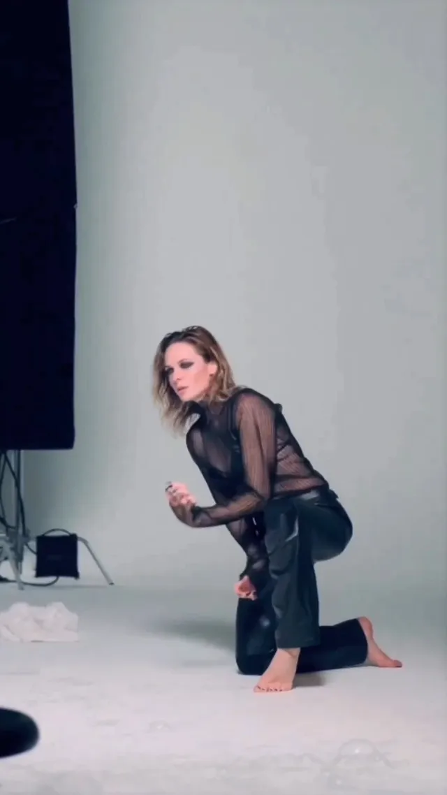 Rebecca Ferguson kneeling barefoot on a studio floor in a dark sheer outfit.