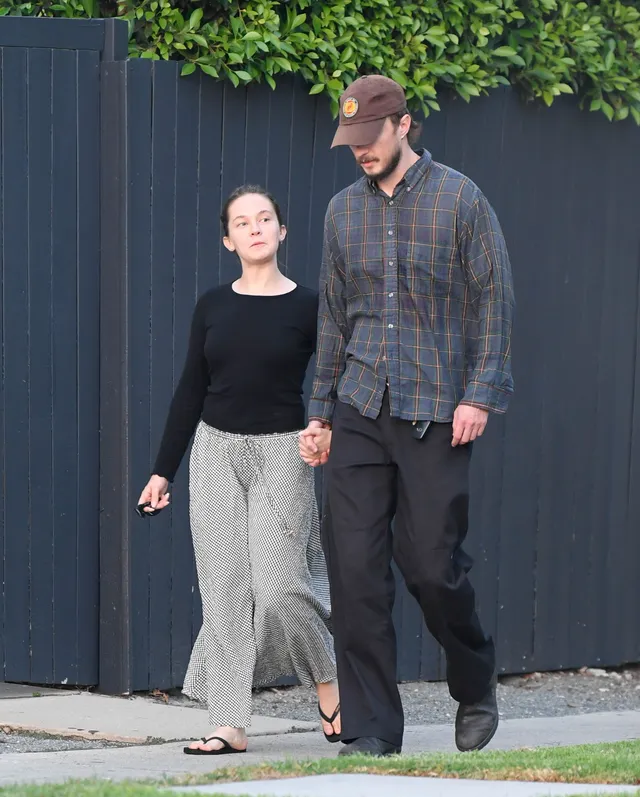 Cailee Spaeny walking on grass in a black top, trousers and flip-flops.