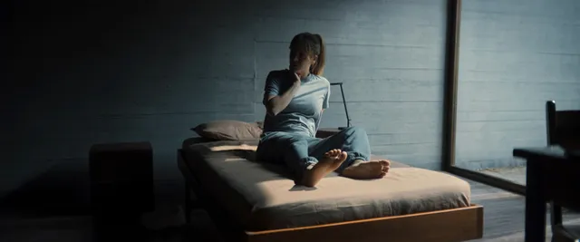 Kaley Cuoco sitting barefoot on a bed in a dimly lit film scene.