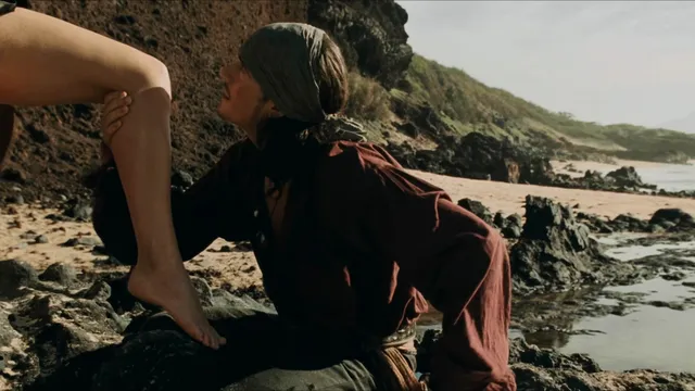 Keira Knightley a seaside screen scene seated among rocks.