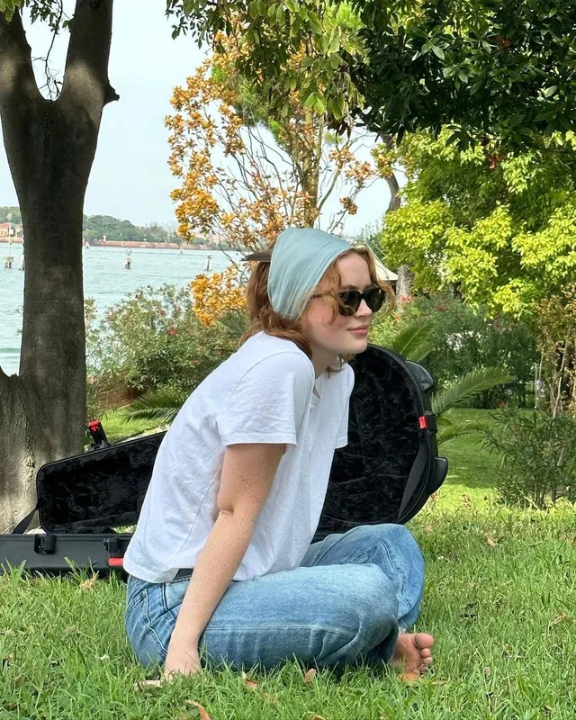 Sadie Sink sitting barefoot on grass in jeans.