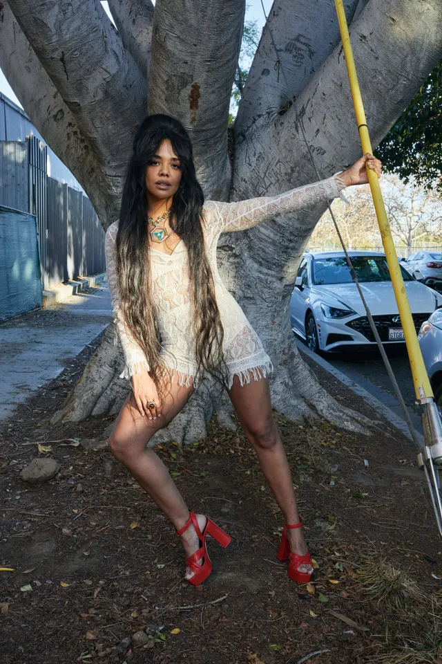 Tessa Thompson posing outdoors in a pale dress and red boots.