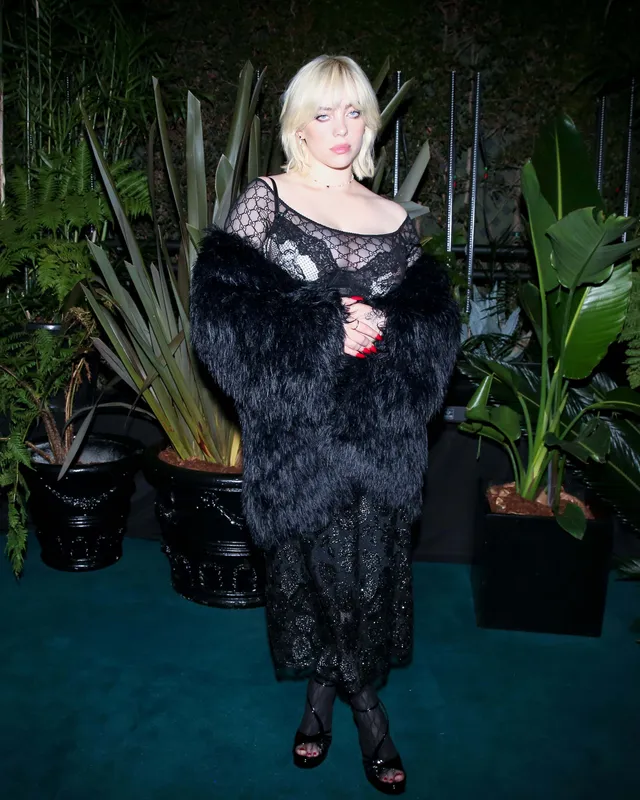 Billie Eilish standing beside plants in a black lace dress and heels.