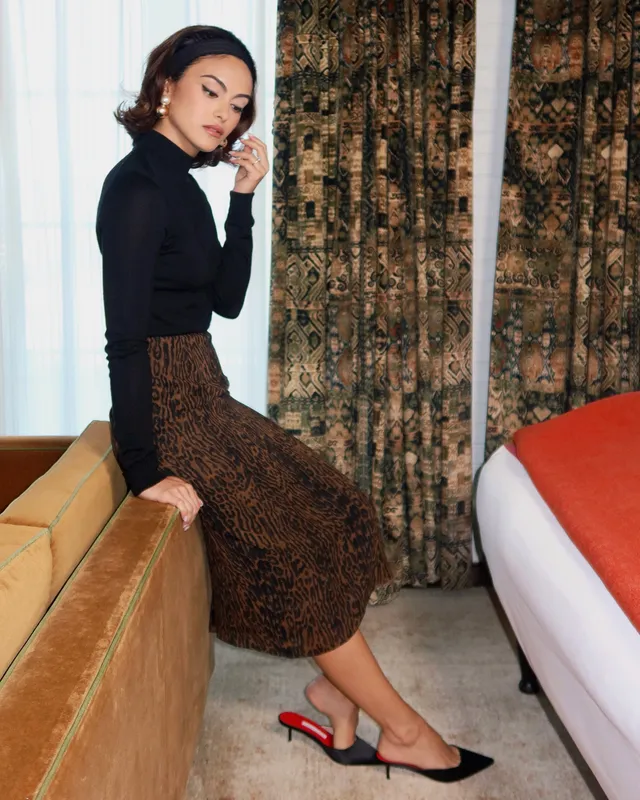 Camila Mendes sitting on a counter in a patterned skirt and black heels.