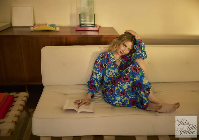 Emma Roberts sitting barefoot on a white sofa in a colorful suit.