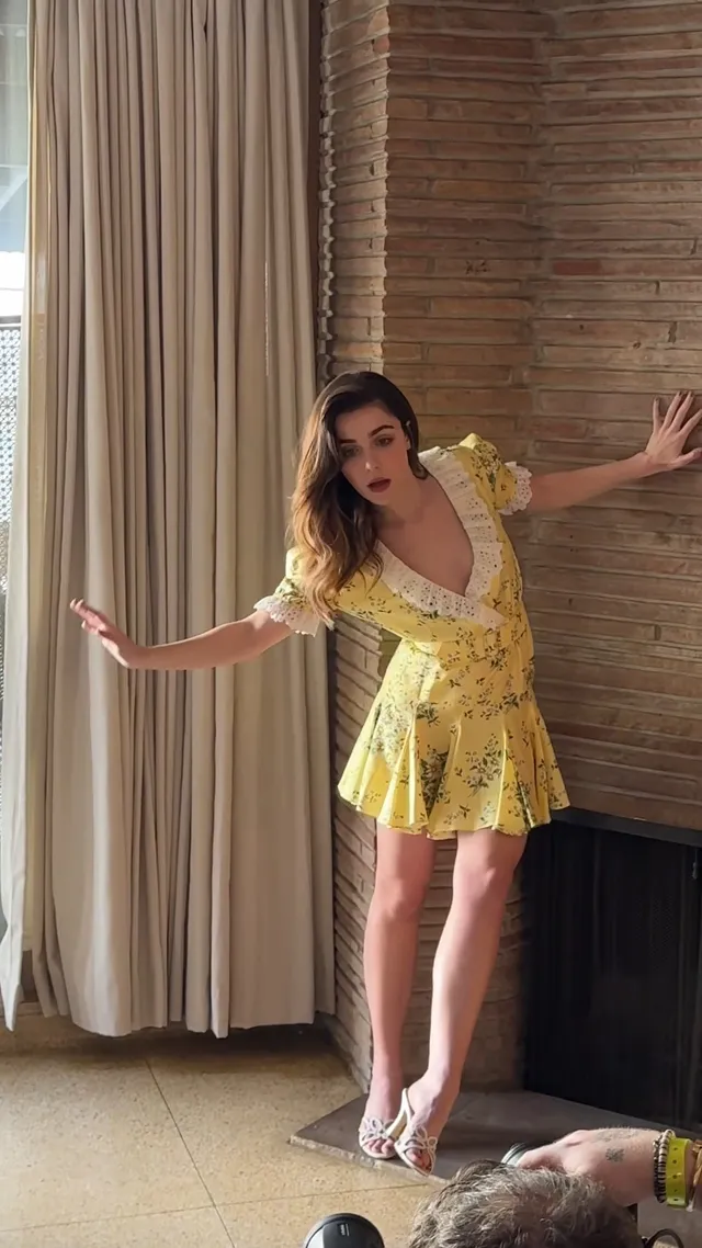 Kiernan Shipka posing by a fireplace in a yellow dress and white heels.