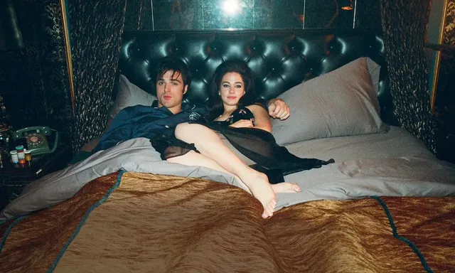 Cailee Spaeny reclining barefoot beside a man on a bed.