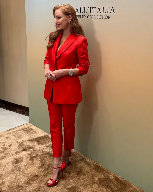 Jessica Chastain standing indoors in a red suit and sandals.