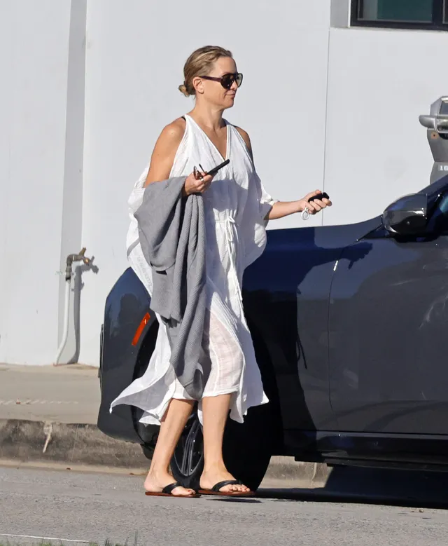 Kate Hudson walking outside in a white dress and flat sandals.